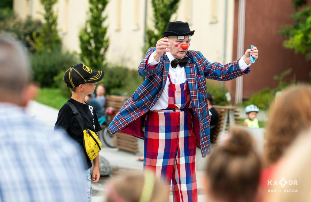 Klaun w meloniku, kraciastych spodniach i marynarce w kratkę przemawia do dzieci na placu